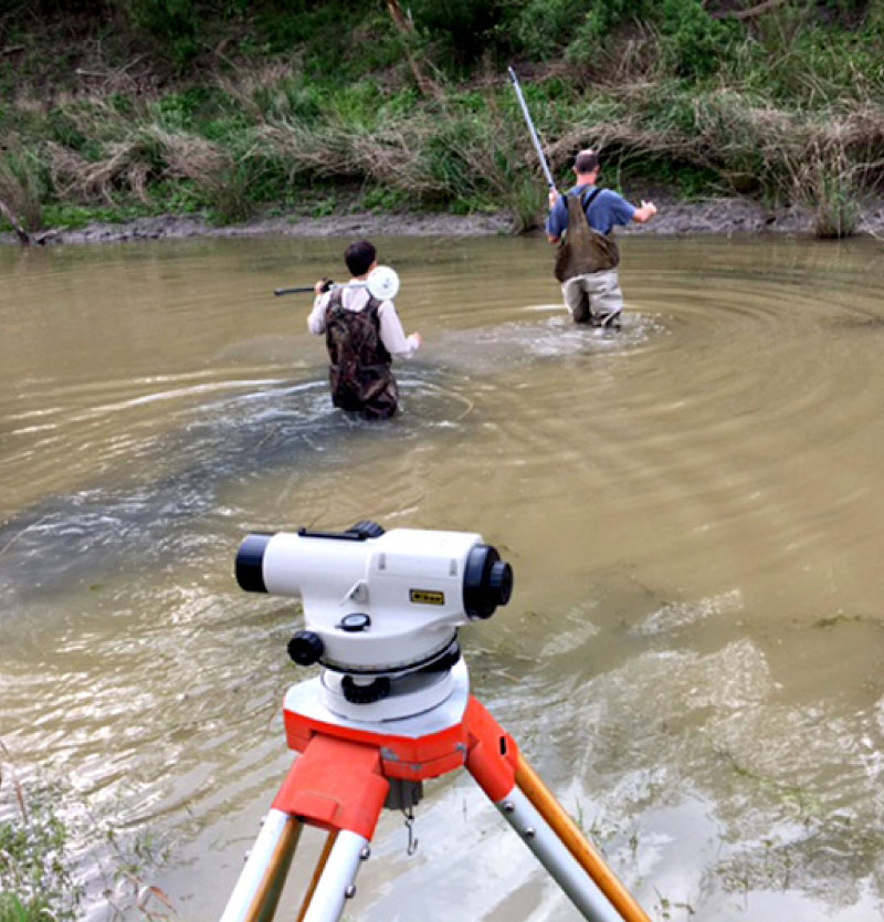 Land surveyors wading through TX stream with surveying equipment